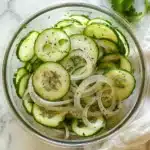Tasty-style overhead photo of cucumber and onion salad in a glass bowl on a white surface with soft natural lighting.