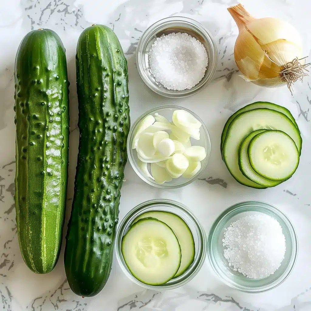 Overhead photo of fresh cucumbers, sweet onion, vinegar, water, salt, and sugar arranged on a marble surface.