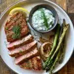 Tuna steak served with roasted asparagus, onions, lemon wedge, and yogurt sauce on a white plate.