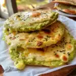 Golden avocado flatbreads with melted cheese and sesame seeds stacked on a parchment-lined white plate, served on a wooden board in natural light.