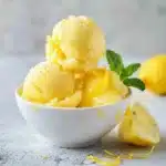 Creamy homemade lemonade sorbet with lemon zest and mint in a white bowl, slightly melting on a linen cloth